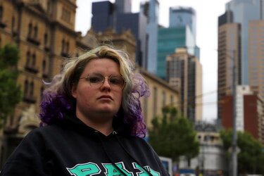 A person with blonde and purple hair with wire-framed glasses in a black jumper and CBD buildings in the background A person with blonde and purple hair with wire-framed glasses in a black jumper and CBD buildings in the background