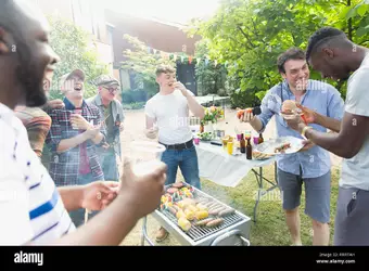 male-friends-laughing-and-eating-around-barbecue-grill-in-backyard-RRRTAH.jpg